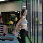 A young woman stretching inside a modern gym with large windows, focused on fitness.